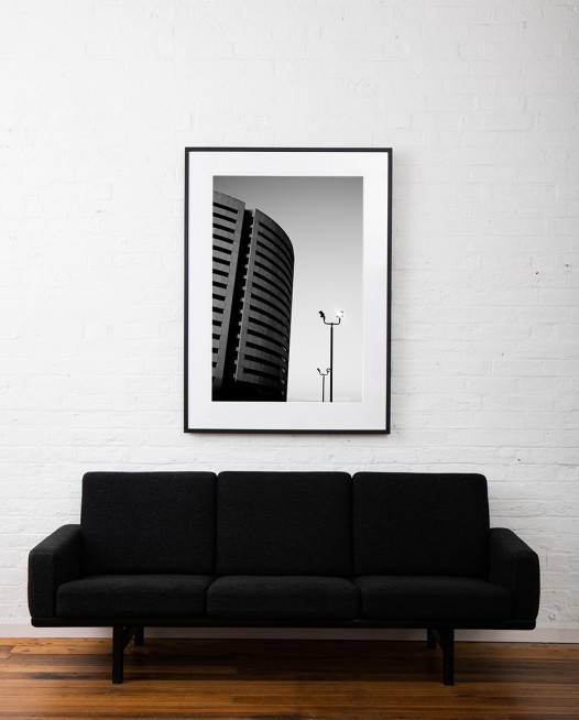 A large Vertical black and white photographic art print of a building and street lamps framed in black timber on wall above sofa