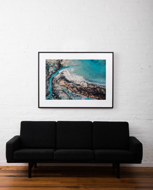 A horizontal abstract aerial photo of water taken in Australia in colour of blue, green and grey framed in black timber on white wall above sofa