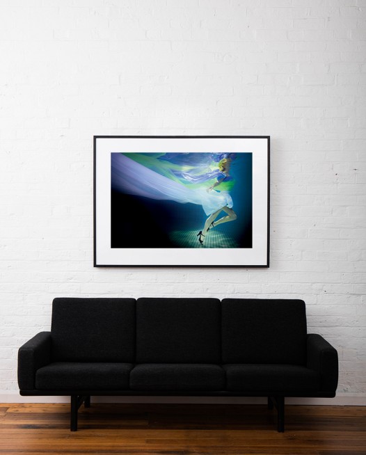 A large photographic art print of a girl under water in blue, green and purple framed in black timber on white wall above sofa