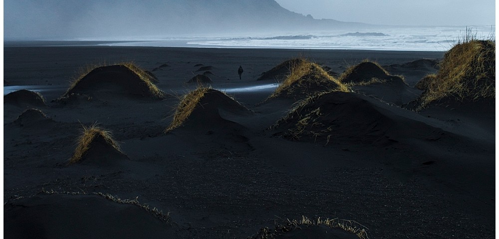 A Icelandic landscape art photograph of sea and beaches with misty mountains background. there is lone person walking on the beach