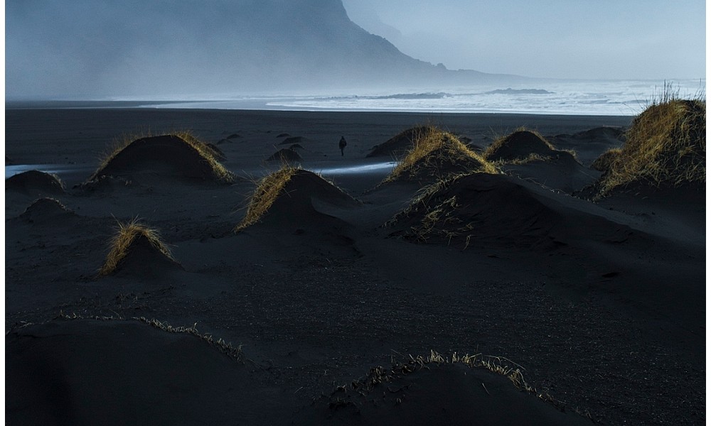 A Icelandic landscape art photograph of sea and beaches with misty mountains background. there is lone person walking on the beach