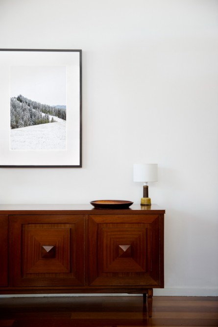 A vertical photographic landscape print of snow, moutain and trees taken in North America in shades of white black and green framed in black timber on wal above cabinet