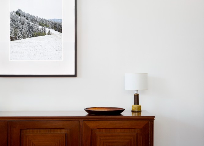 A vertical photographic landscape print of snow, moutain and trees taken in North America in shades of white black and green framed in black timber on wal above cabinet