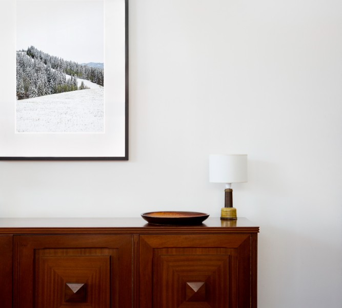 A vertical photographic landscape print of snow, moutain and trees taken in North America in shades of white black and green framed in black timber on wal above cabinet