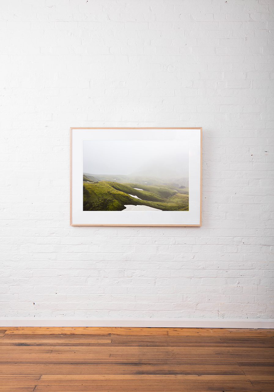 A Large photographic Image of misty green Iclandic landscape of mountains and snow framed in raw timber on white wall