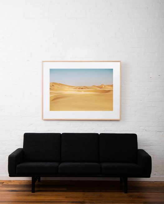 A photograph of Africa desert in yellow sand and blue sky framed with raw timber on white wall above sofa