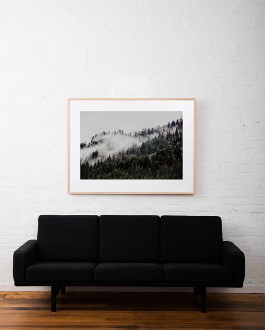 A photographic print of mist and fog over Mountains and trees in Canada framed in raw timber on white wall above sofa